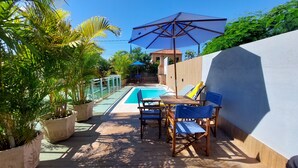 A pool area with a table and chairs, surrounded by potted plants and a blue umbrella. - Village Itaúna Inn – Beach Houses (Saquarema)