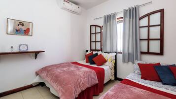 A bedroom with two beds, a shelf with books, a painting, and an air conditioning unit.