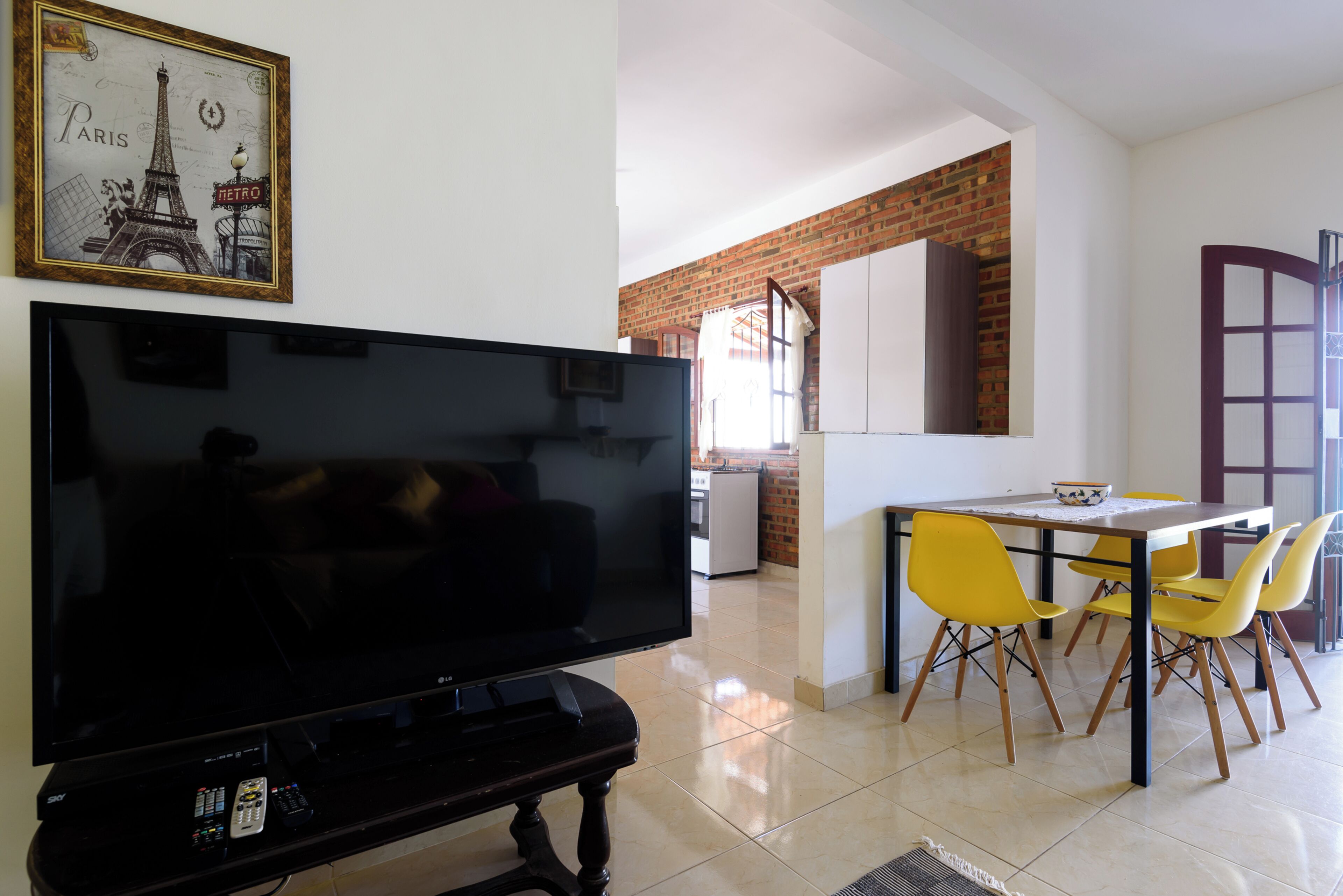A living room with a flat-screen TV, a dining area with yellow chairs, and a kitchen in the background.
