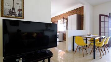 A living room with a flat-screen TV, a dining area with yellow chairs, and a kitchen in the background.