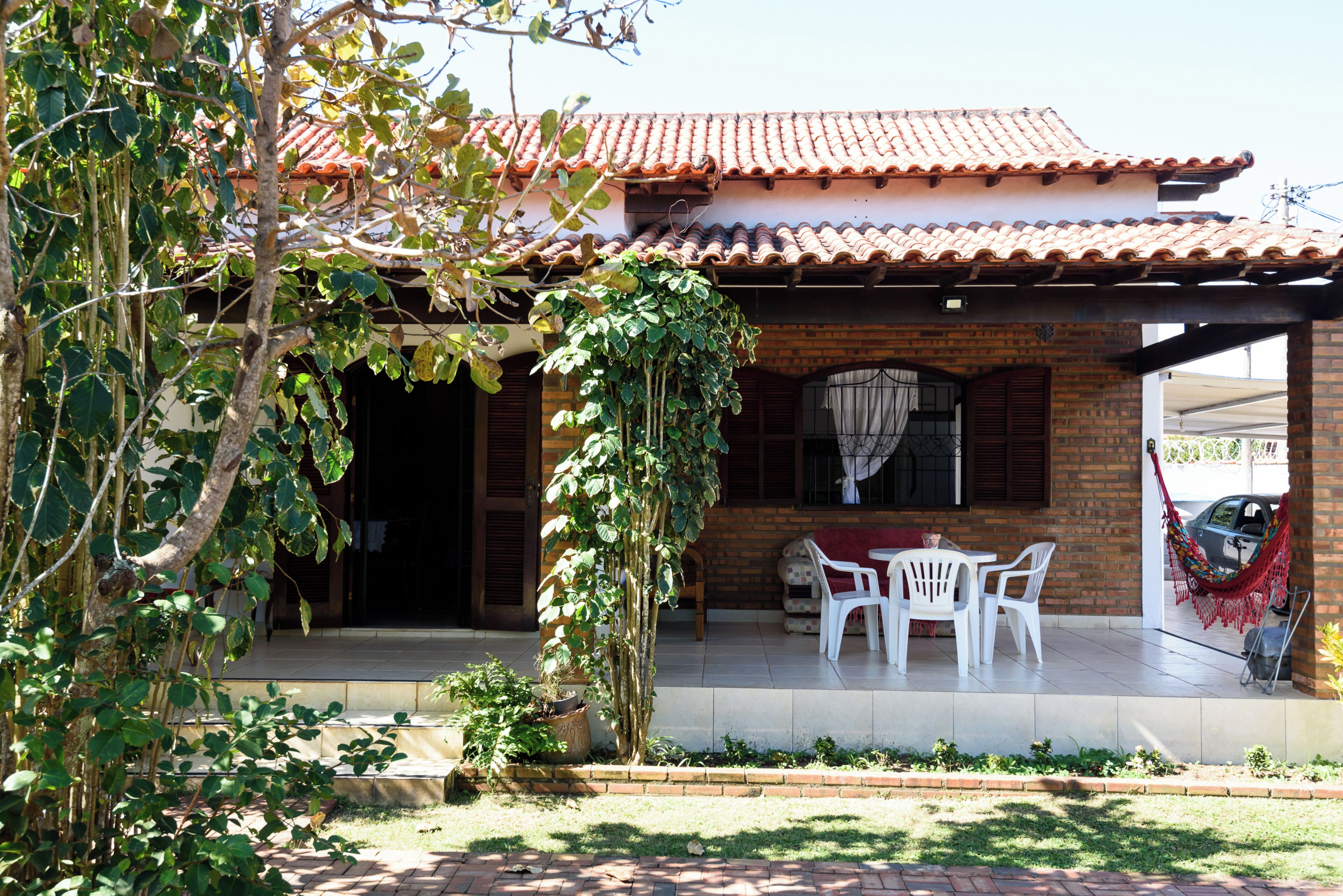 Una casa con techo de tejas, fachada de ladrillo y un patio con sillas blancas de plástico.