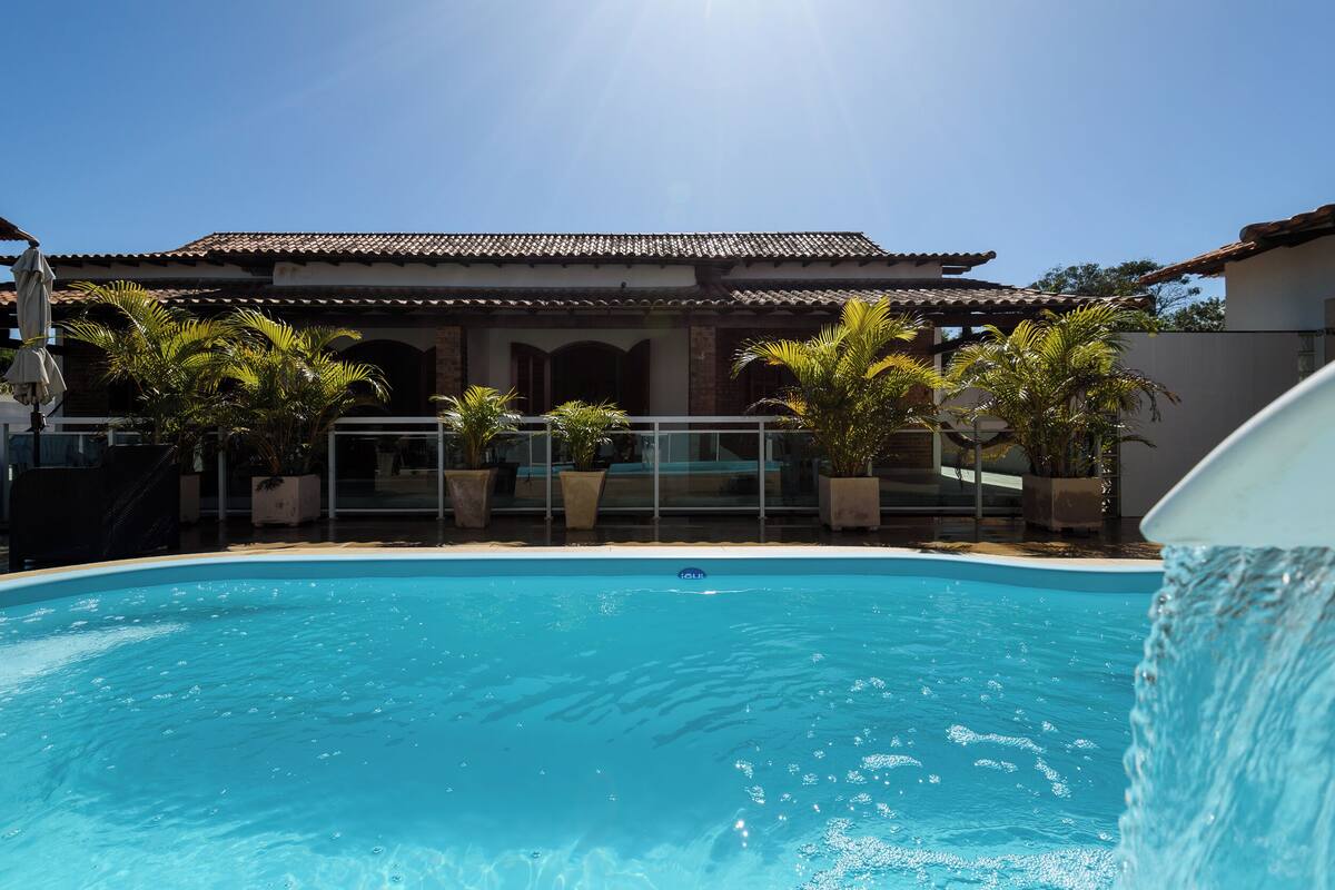 Una piscina con agua cristalina y azul, rodeada de macetas con plantas y un edificio con tejado de azulejos.