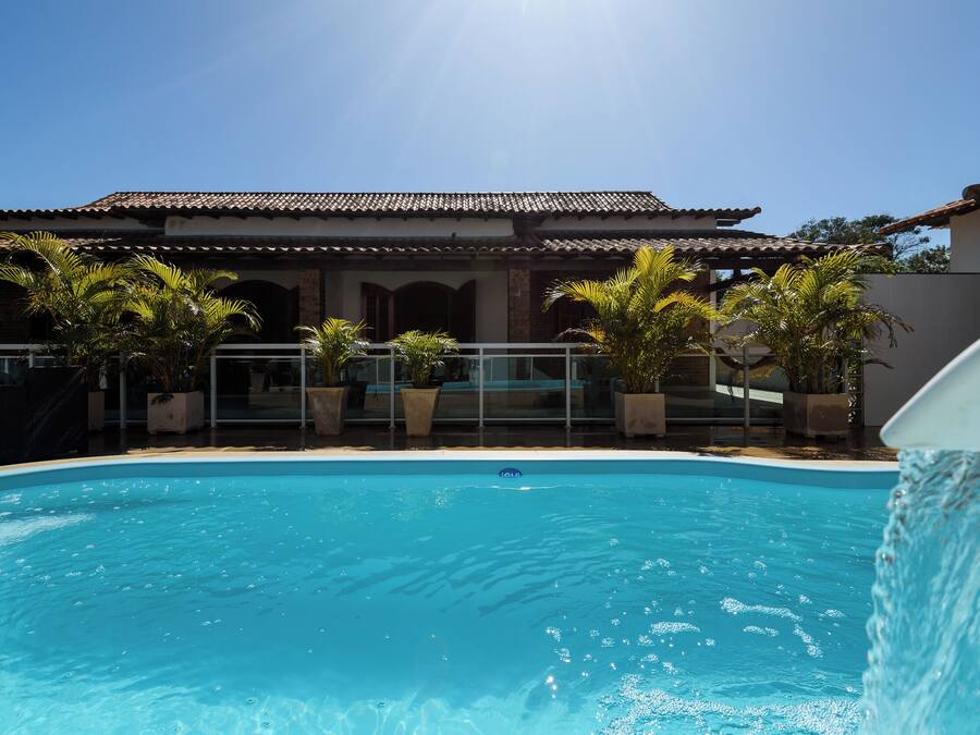 Une piscine à l’eau d’un bleu limpide, entourée de plantes en pot et bordée d’un bâtiment dont le toit est recouvert de tuiles.