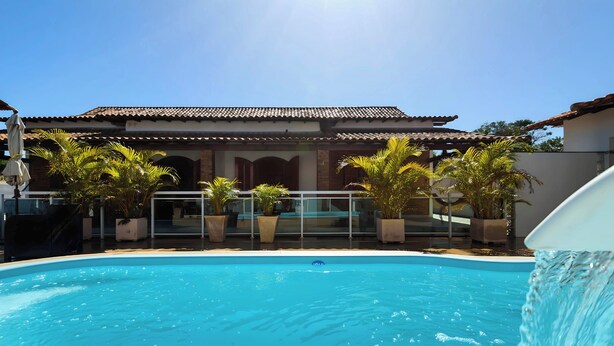 A swimming pool with a clear blue water, surrounded by potted plants and a building with a tiled roof. - Village Itaúna Inn – Beach Houses (Saquarema)