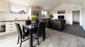 A modern kitchen with a dining area, a living room with a sofa and TV, and a large window. - NV Homes (Lake Hayes)