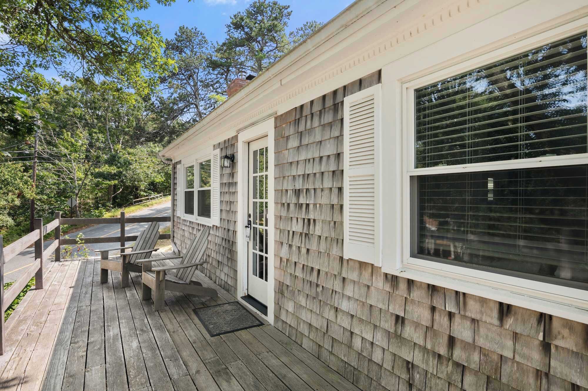 A wooden deck with two Adirondack chairs and a bench, a covered porch, and a building with shingled siding and white trim.