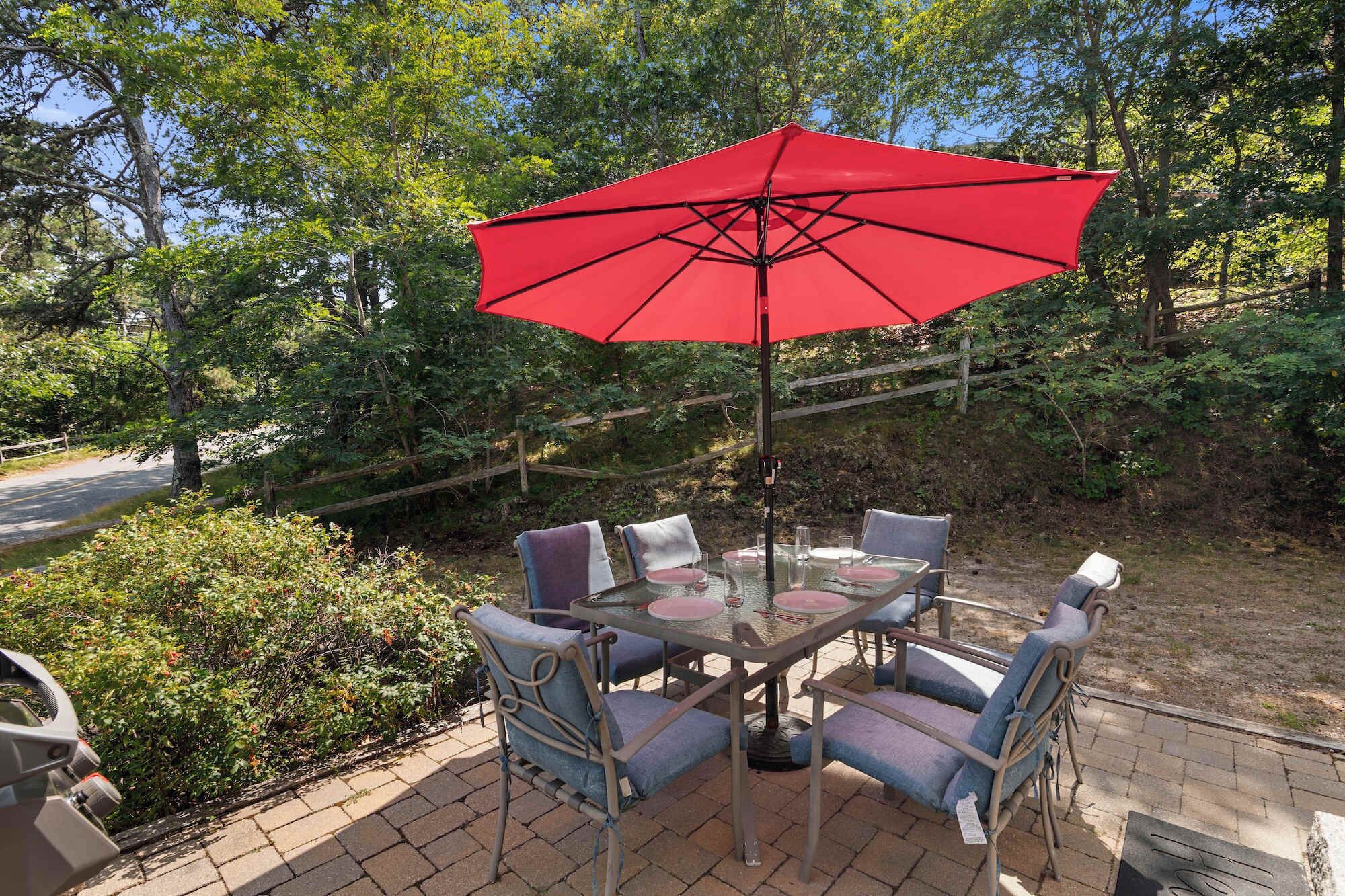 A patio with a table and chairs under a red umbrella, surrounded by greenery.