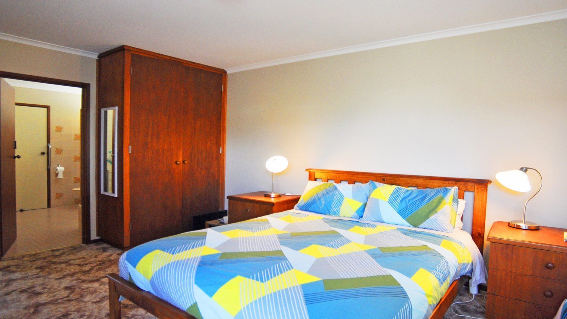A hotel room with a large bed, wooden headboard, two bedside lamps, a wooden wardrobe, and a door leading to a bathroom.