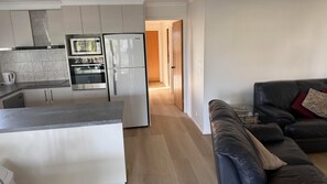 A modern kitchen with white cabinets, a microwave, and a refrigerator. There is a wooden floor leading to a living area with a black leather sofa and a coffee table.