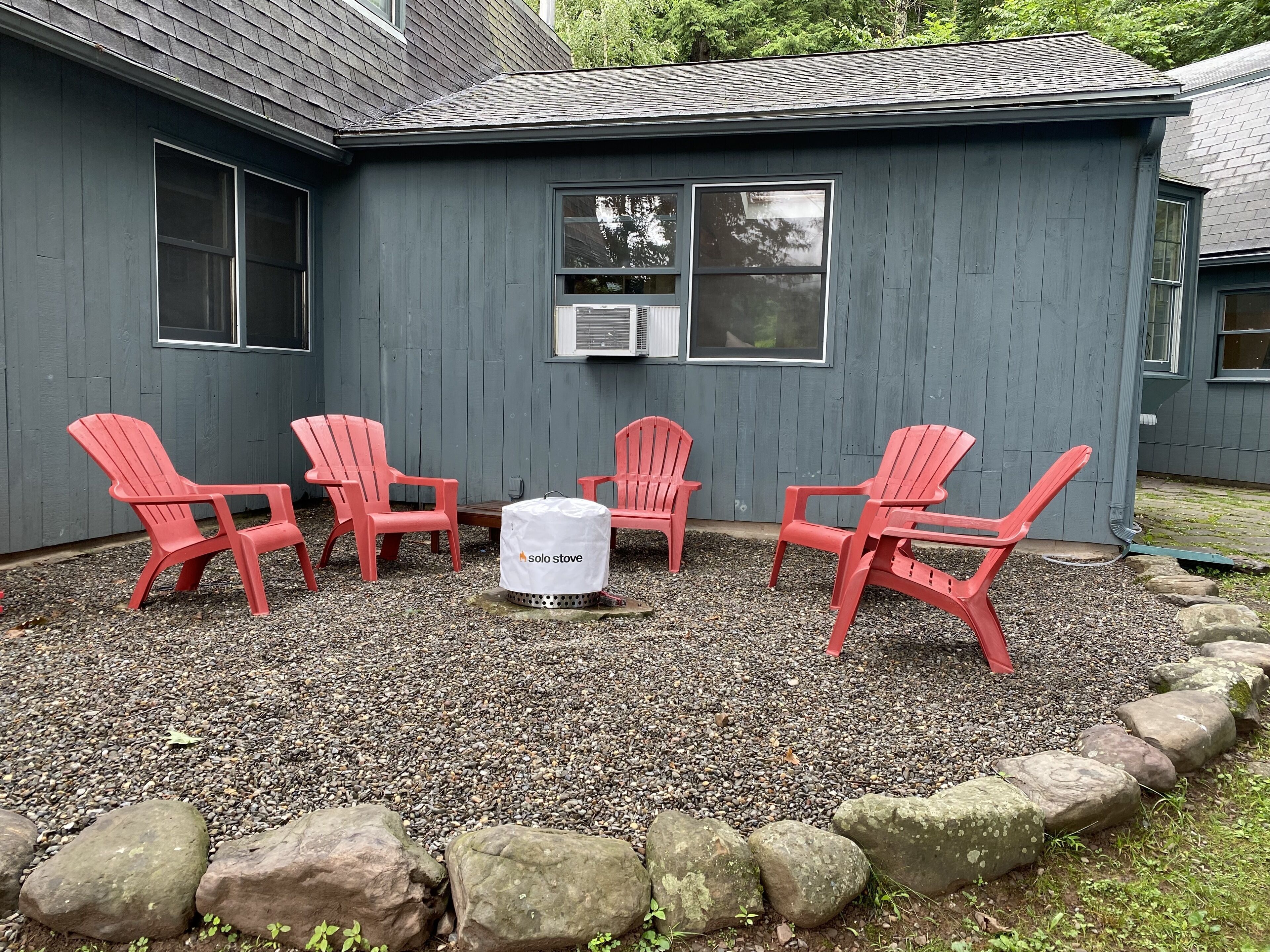 A patio area with four red Adirondack chairs and a white outdoor heater labeled 'solo stove' against a gray building with a window and an air conditioning unit.