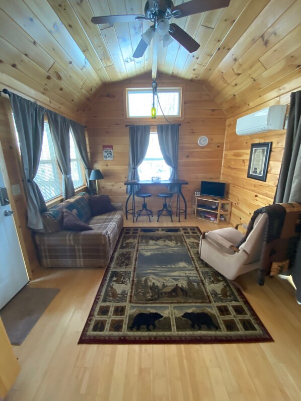L’intérieur chaleureux d’un chalet avec un plafond en bois, un ventilateur de plafond, un canapé à carreaux, une petite table à manger avec deux chaises, un tapis orné de silhouettes d’ours et une fenêtre avec des rideaux.