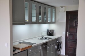 Uma cozinha compacta com armários cinzentos, cafeteira, máquina de lavar e um balcão pequeno.