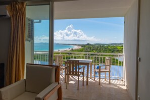 Varanda com mesa e cadeiras, vista para uma praia e paisagem verde.