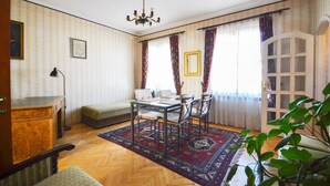 A room with a bed, a desk, a dining table with chairs, a chandelier, and a large window with curtains.