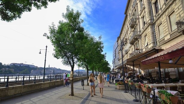 A riverside walkway with outdoor seating, trees, and a building with a sign reading "DINACORSO".