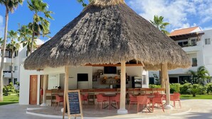 Eine Bar im Freien mit einem Reetdach, Tischen und Stühlen, umgeben von Palmen und einem Gebäude mit Balkonen.
