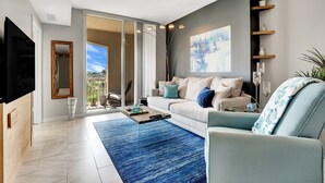 Un salon moderne avec un canapé, une table basse et un téléviseur. Une grande fenêtre offre une vue sur l’extérieur.