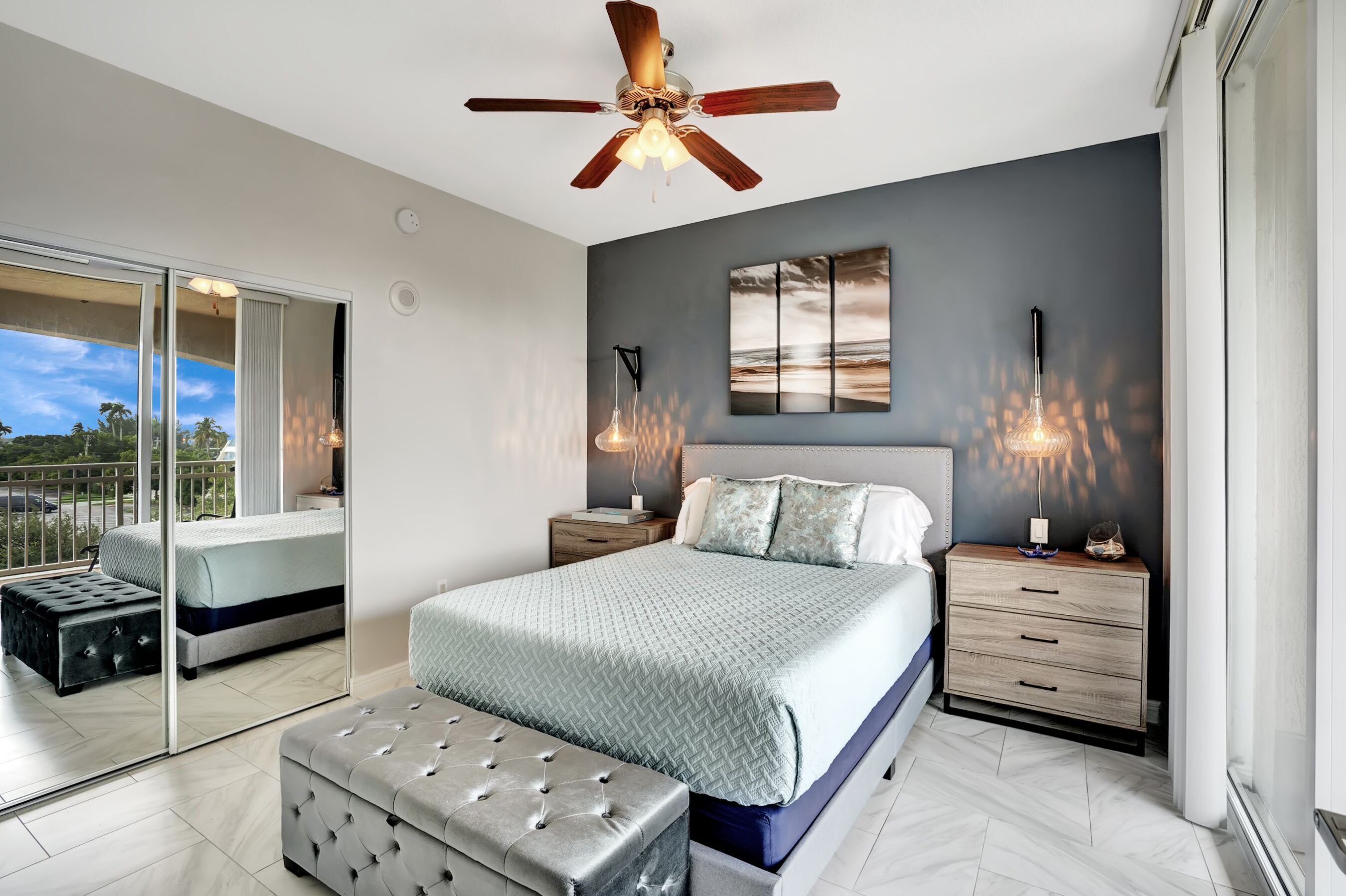 A modern bedroom with a large bed, a ceiling fan, bedside lamps, and a view through a sliding glass door.