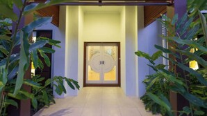 Un pasillo con puerta doble, flanqueado por plantas, que conduce a un edificio de diseño moderno y techo de madera.