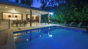 Un área de piscina moderna con una zona de estar cubierta al aire libre, una piscina y exuberante follaje de fondo.