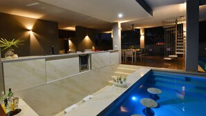 Una cocina moderna con barra integrada, un área de piscina con asientos de descanso y una escalera que lleva a un balcón.