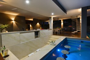 A modern kitchen with a built-in bar, a pool area with lounge seating, and a staircase leading to a balcony.