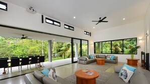 Amplia sala de estar con un sofá grande, una mesa de centro y un comedor con sillas. Amplias ventanas ofrecen vistas a exuberante follaje.