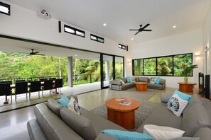 A spacious living room with a large sofa, a coffee table, and a dining area with chairs. There are large windows offering a view of lush greenery.