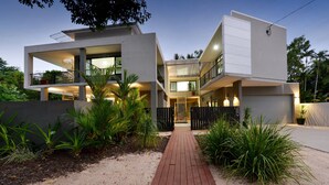 Una casa moderna de dos pisos con una escalera central, rodeada de plantas tropicales y un sendero pavimentado.