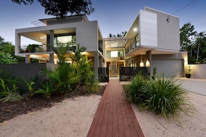 A modern two-story house with a central staircase, surrounded by tropical plants and a paved walkway.