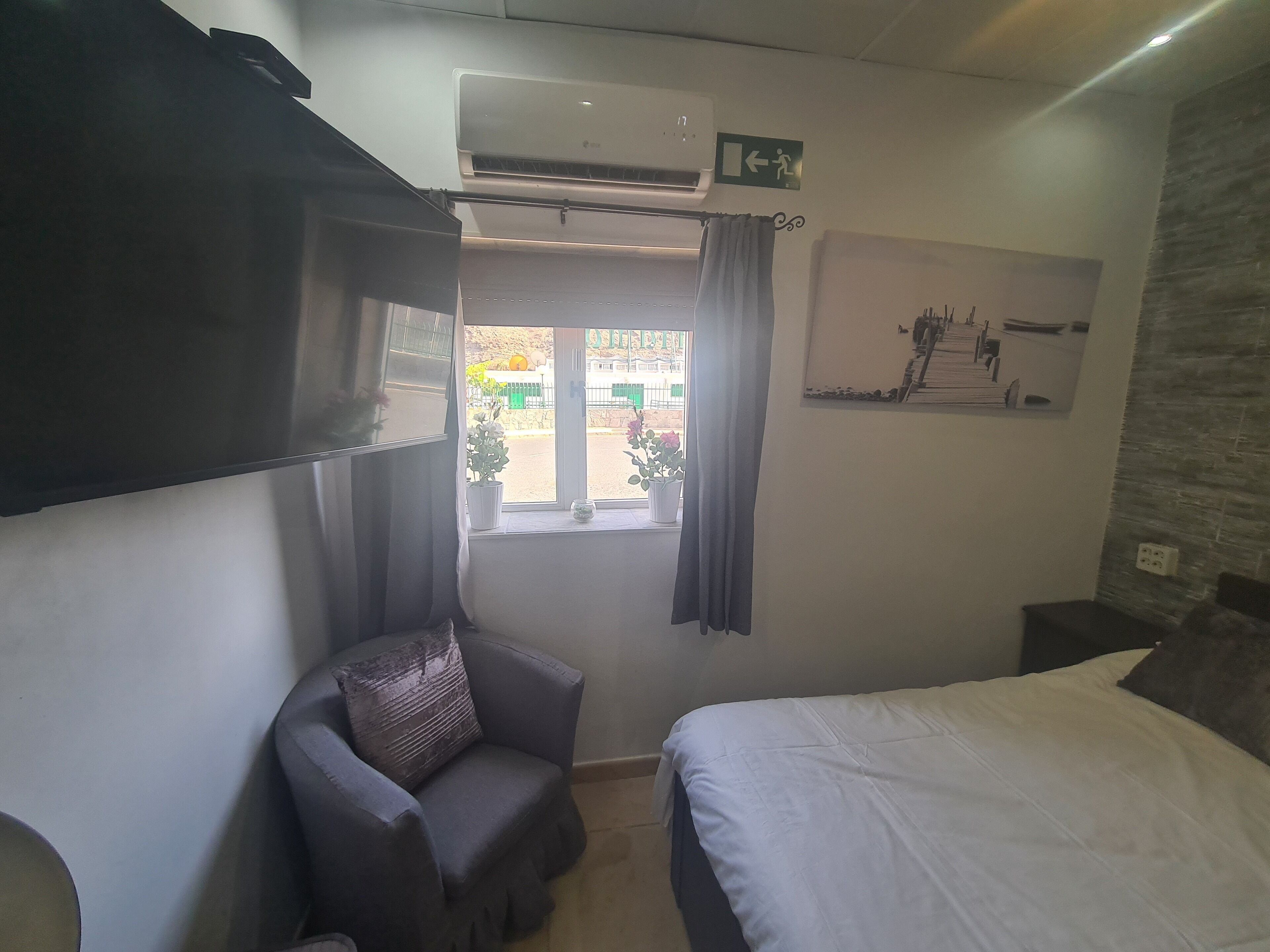 Un dormitorio con cama, una silla, un equipo de aire acondicionado, un ventanal con cortinas y un cuadro colgado en la pared.