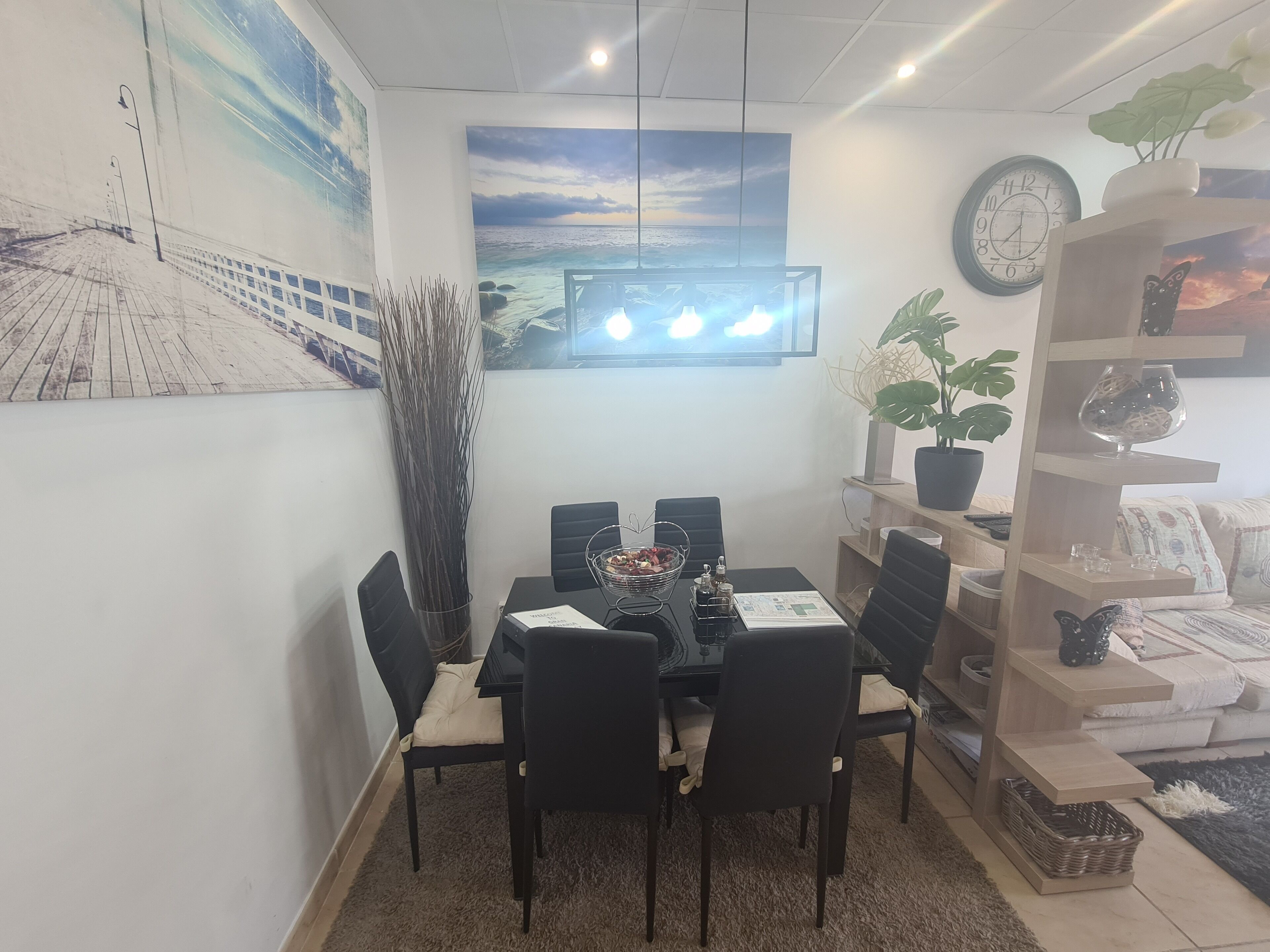 Un comedor con una mesa dispuesta para cuatro personas, rodeada de sillas, un reloj en la pared y un ventanal con vistas al mar.