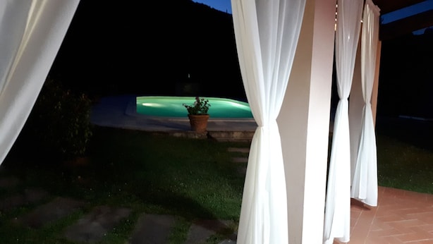 Een overdekt buitengebied met witte gordijnen, een potplant en uitzicht op een verlicht zwembad.