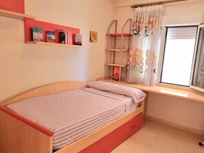 A bedroom with a single bed, a shelf with books, a window with curtains, and a bedside table with a lamp.