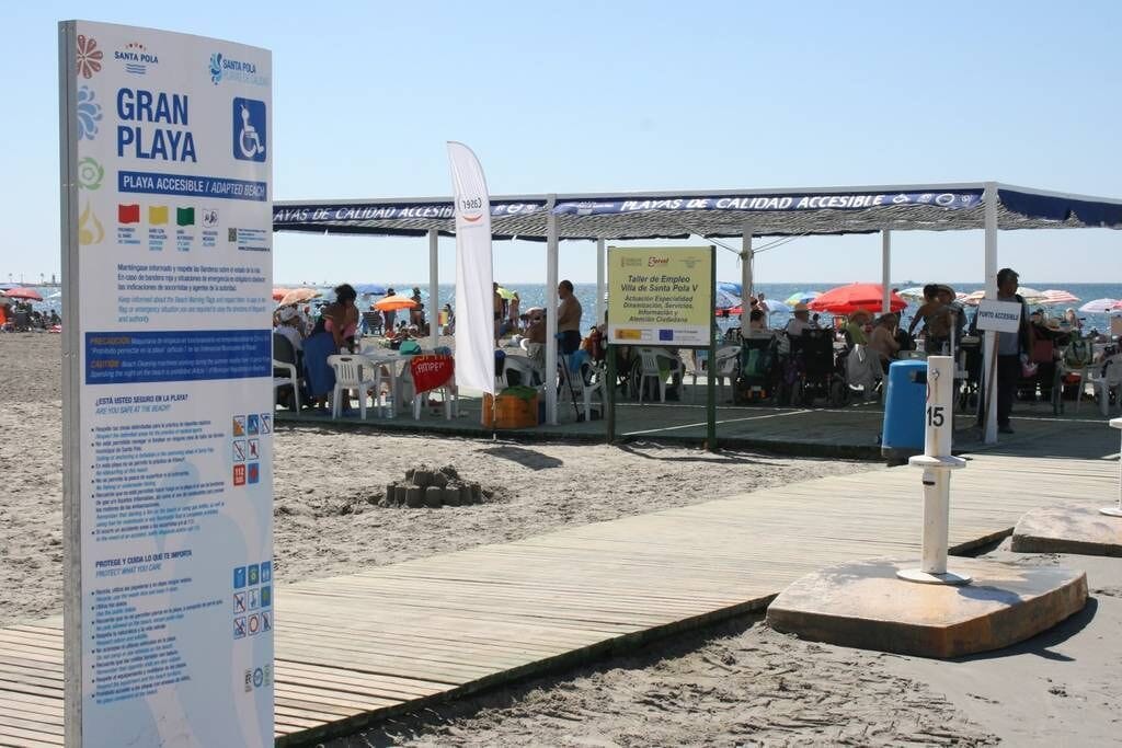Une zone de plage avec un panneau indiquant « Gran Playa » et « Playa Accesible / Plage aménagée pour les personnes à mobilité réduite ». On aperçoit des parasols et des personnes au loin.