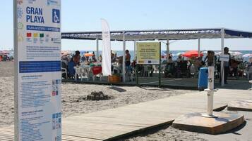 Une zone de plage avec un panneau indiquant « Gran Playa » et « Playa Accesible / Plage aménagée pour les personnes à mobilité réduite ». On aperçoit des parasols et des personnes au loin.