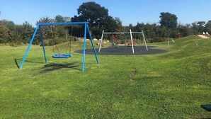 Un parque infantil con columpios y tobogán, rodeado de césped y árboles.