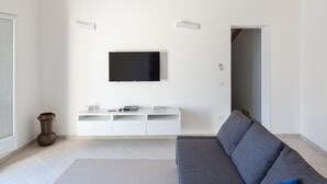 A modern living room with a grey sofa, a flat-screen TV mounted on a white wall, and exposed wooden beams on the ceiling. - Attico Mansion, Ortigia (Siracusa)