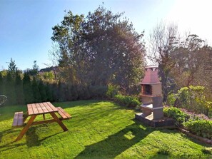 A wooden picnic table and a stone barbecue grill in a grassy garden with trees in the background. - Apartment 2: With porch, pool and garden (Tapia De Casariego)