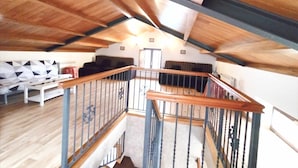 A spacious loft with a high ceiling, wooden beams, a staircase with a wooden handrail, and a seating area with a sofa and a bench.