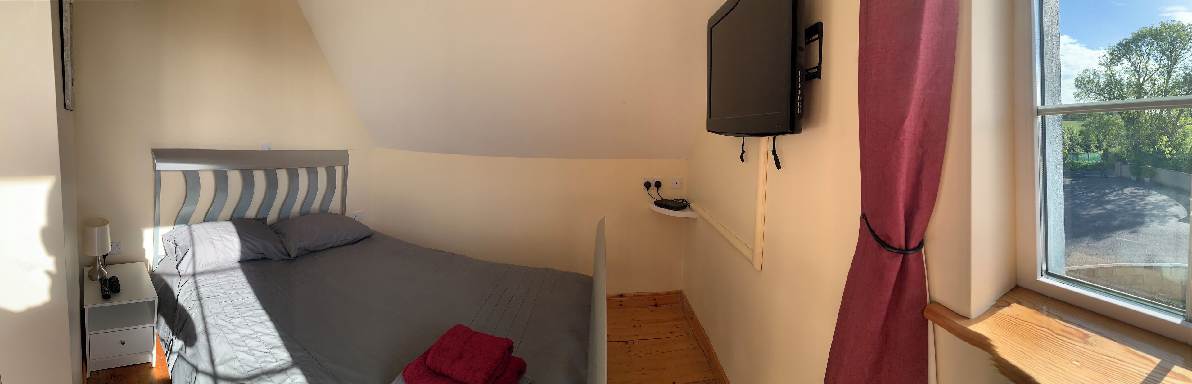 Un dormitorio con cama, mesita de noche con lámpara y control remoto, televisor montado en la pared y ventana con vistas al exterior.