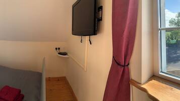 Un dormitorio con cama, mesita de noche con lámpara y control remoto, televisor montado en la pared y ventana con vistas al exterior.