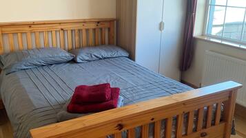 A bedroom with a wooden bed, grey bedding, a red towel, a wardrobe, and a window with curtains.
