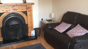 Un salón con chimenea, un sofá de cuero negro, una mesita con una lámpara y un televisor montado en la pared.