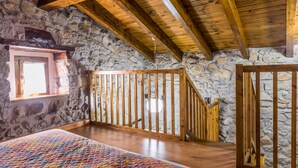 A cozy room with a stone wall, wooden ceiling beams, a colorful quilted bedspread, and a window with a view.