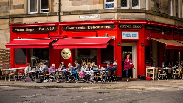 An outdoor café with tables and chairs, a red awning, and a sign reading "Victor Hugo Est 1940 Continental Delicatessen." - Main Door Apartment in an enviable location (Edinburgh)