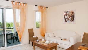 Una sala de estar con un sofá blanco, una mesa de centro de madera y una mesa de comedor. Hay dos sillas de mimbre, un ventanal que da al exterior y un cuadro en la pared.