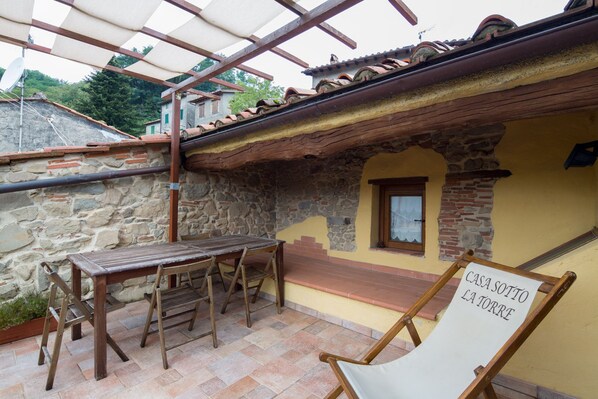 A rustic stone building with a wooden table and chairs, a sign reading "CASA SOTTO LA TORRE", and a staircase leading to a window.