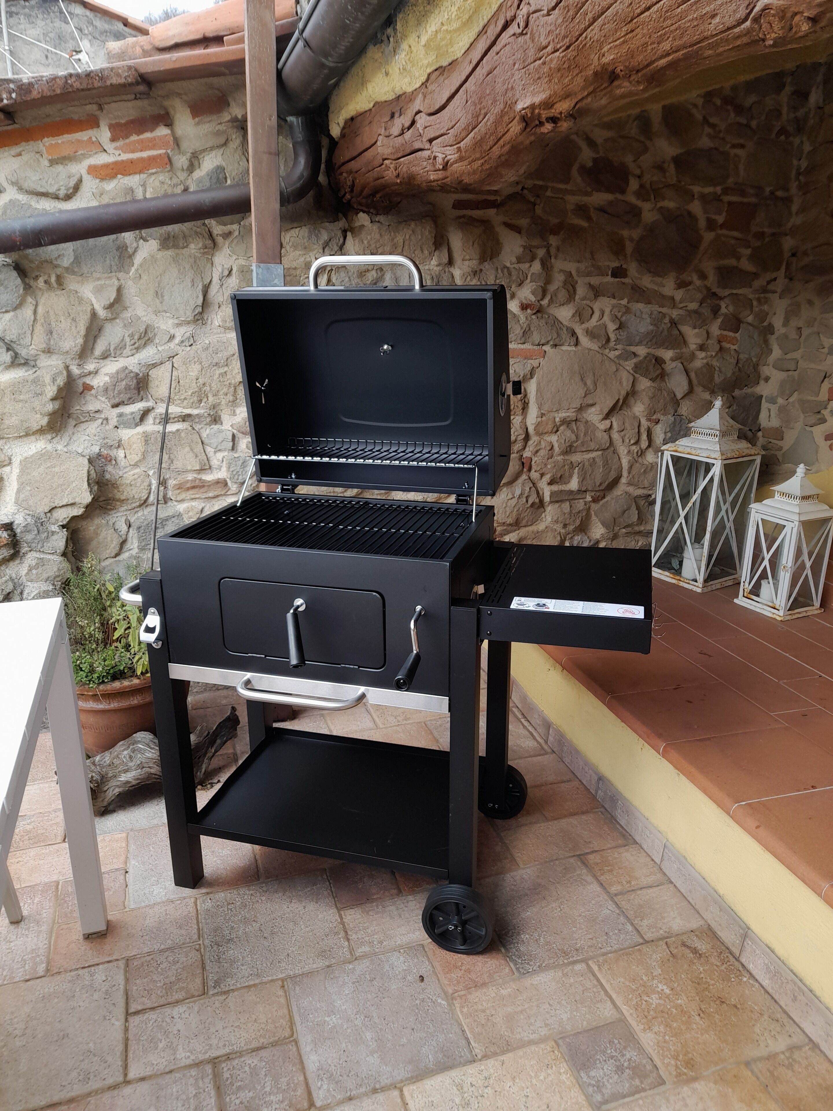 A portable grill with wheels, set on a patio with stone walls and a tiled floor.