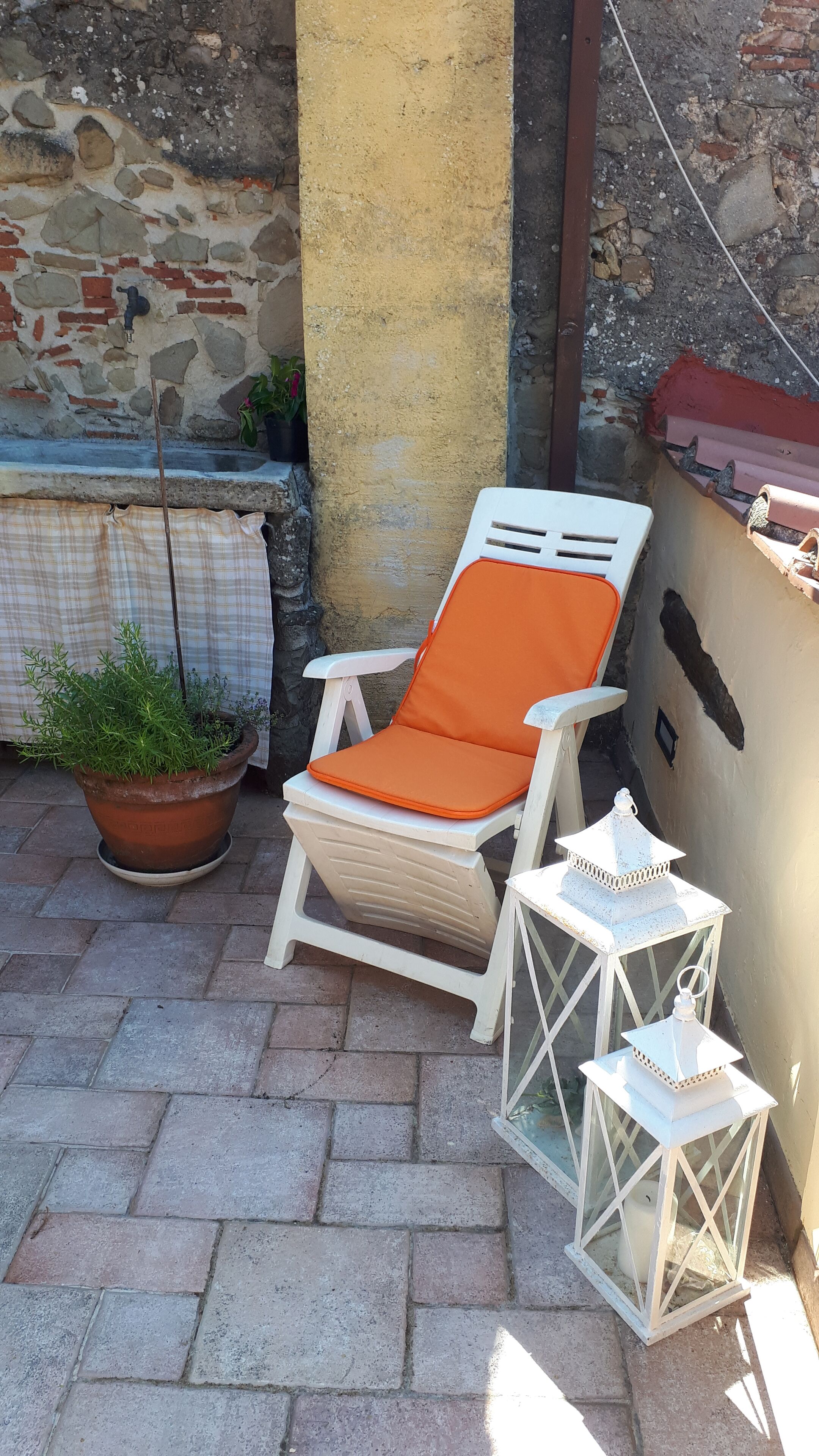 Un fauteuil de jardin blanc avec un coussin orange, une plante en pot et deux lanternes blanches sur une terrasse dallée.